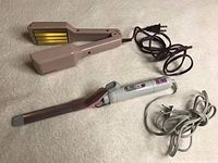 Two electric hair styling tools: beige zig-zag crimper and rose gold/white curling wand with clamp, cords coiled, placed on light fabric surface.