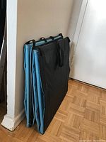 Two folded beach recliners standing upright against a wall. They have black fabric backing and black carrying handles with light blue padded seats hidden inside.