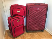 Three pieces of luggage: large red and black checkerboard patterned suitcase with Via Rail logo, two smaller red soft-sided bags with matching logos, arranged against a wall.