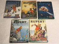 Five Rupert annual books arranged on a flat surface, showing colorful illustrated front covers featuring Rupert Bear in various scenes such as skating, fishing, space, riverbank, and hill climbing.