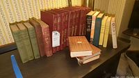 Photo showing thirteen antique books on a shelf of various sizes and binding colors including red, green, and blue. Books show wear and some damage.