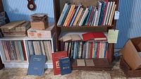 Wooden bookshelf with three shelves filled with various books, and additional books in a white side shelf and stacked on the floor