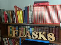 Shelf with a variety of books including hardcovers of various colors and a set of red and white Readers Digest Man & Woman volumes with wooden letters spelling 'FLASKS'.