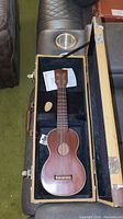 Open tweed hard case holding mahogany soprano ukulele, pick and string package visible