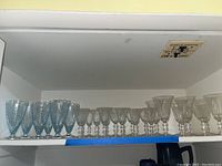 Top shelf holding an assortment of stemware glasses including six blue crystal water glasses and clear glass stemware glasses all with etched patterns.