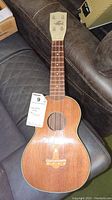 Full front view of ukulele showing body, neck, headstock, and attached lot tag
