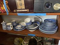 Shelf displaying Lamberton blue and white transferware dishes including stacked plates, cups, pitchers, a covered pot, and bowls.