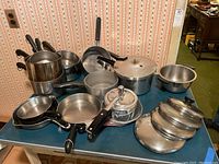 Wide view of various pots, pans, lids, and pressure cookers arranged on a table against wallpaper background