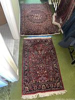 Two area rugs shown side-by-side on green carpet, displaying their overall size and patterns. The smaller rug has a red base with floral designs. The larger rug shows a pattern with a central octagon.
