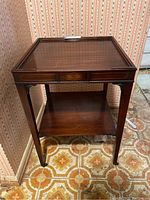 Front and side view of a wooden side table with glass top and decorative inlaid oval on the front apron, showing scratches and casters at the bottom.