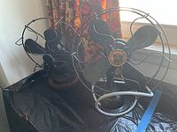 Two black vintage electric fans displayed side by side on a table near a window with floral curtains.