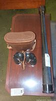 Photo showing the full lot on a wooden table: the spyglass, binoculars, and leather case.