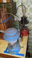 Photo showing three vintage lanterns together. Front lantern is blue Coleman with Sears branding, middle is a second blue Coleman lantern, back is red Kamplite lantern missing glass globe.