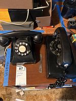 Photo showing two black rotary dial telephones, one wall-mounted Bell System Western Electric phone and one vintage table phone on a wooden surface with various items around.