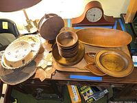 Various wooden salad bowls, a large oval bowl, round bowls stacked, divided serving tray, and woven rattan baskets arranged on wooden furniture surface under lighting. Mantle clock visible but not included.