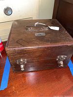 Closed wooden box with metal latches and handle, showing external wear and attached metal identification plate.