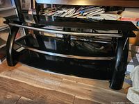 Full view of the black TV stand showing top and middle glass shelves with chrome support rods and curved legs.