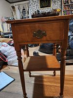 Front view of the Ethan Allen night table showing drawer, turned legs, and lower shelf on wooden floor with furniture and bags in background.