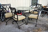 Full view of round glass top table surrounded by four metal frame chairs with cream-colored cushions, showing the overall set and chair styling with castor wheels on chair legs.