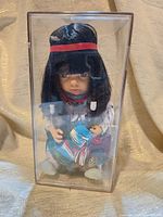 Front view of Native American baby doll in clear display case showing black straight hair with red headband and traditional dress with fringe and bead details.