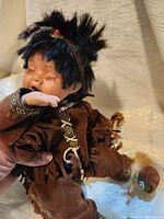 Front and side view of the Native American porcelain doll seated and wearing brown suede fringe outfit with beadwork.