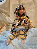 Front view of the Native American porcelain doll sitting, showing detailed facial features, traditional costume, and accessories including feathers and a small drum.