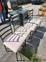 Front angled view of four vintage metal chairs showing black ladder-back frames and light-colored upholstered seats, set outdoors on concrete surface.