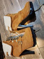 Side view of tan suede ankle boots with shearling lining, studded trim, lace-up front, buckle strap and high stacked heel on wooden floor.