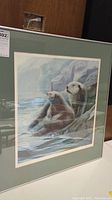 Front view of framed artwork showing two sea otters sitting on rocks near water, with a green mat and glass reflection.