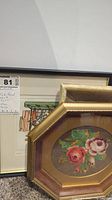 Photo showing two petit point floral artworks with gold-toned octagonal frames with a matte border, leaning against a wall.