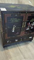 Front angled view of black lacquer Asian themed cabinet showing inlaid figures and landscape on doors, and animal-shaped drawer pulls.