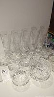 Group of various crystal glasses including tall glasses, coup glasses, and bowls laid out on a table under lighting revealing cut patterns.