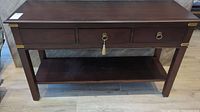 Front view of wooden sofa table with three drawers, one missing a pull, showing dark brown finish and brass corner accents