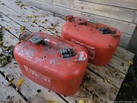 Photo of two orange boat fuel tanks on a wooden surface outdoors with some leaves around. Both tanks show wear and have fuel caps.