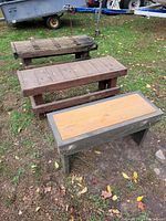 Three wooden benches placed outside on grass. One bench in the back has holes on the top surface and appears worn. The other two benches in front have solid wooden tops with different wood colors.