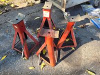 Four red metal jack stands placed on an outdoor concrete surface with visible dirt and leaves.