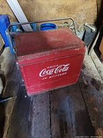 Red metal Coca Cola cooler with white logo and carry handle shown from front angle on wood floor.
