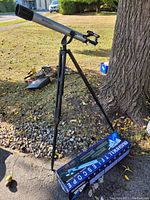 Full side view of Bushnell telescope mounted on black tripod stand outdoors, original box on ground beside tripod