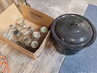 Photo showing several clear glass canning jars with metal lids in a cardboard box next to a large black enameled metal canning pot with lid on.