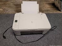 Front view of Canon white all-in-one printer showing paper tray, control panel, and connected power cord on wooden floor.