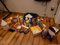 A collection of various cleaning products and tools arranged on a wooden floor including bottles of cleaning solutions, sponges, brushes, and packaged boxes.