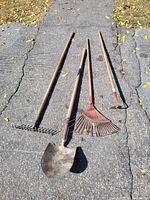 Four garden tools laid on pavement including 2 rakes, 1 shovel, 1 hoe