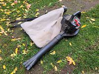 Gas leaf blower laid on grass with attached beige fabric collection bag and black plastic nozzle.