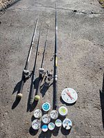 Photo showing four fishing rods aligned on the ground with three reels mounted on rods, with eight extra fishing line spools and a mechanical weigh scale beside them.