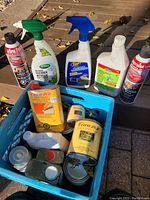 Blue milk crate containing assorted cleaning and paint maintenance products on an outdoor surface.