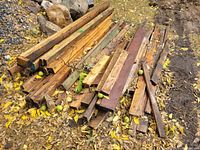 First image showing a pile of various wooden boards and planks of differing lengths and widths, placed on the ground with fallen leaves around.