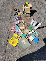 Wide view of all items arranged on pavement including books, adhesives, pacifiers, small vintage collectibles, a photo album, and craft supplies
