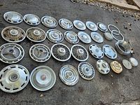 Overall collection of vintage metal hub caps and rings laid out on pavement showing different styles and conditions.