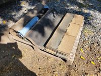 Pallet containing several bundles of asphalt shingles stacked alongside rolls of roofing felt material, placed on outdoor ground surface with gravel and dirt visible, shot in daylight with some shadow from adjacent trees.