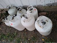 Photo of seven used propane tanks of varying sizes and conditions shown outdoors against a building wall.